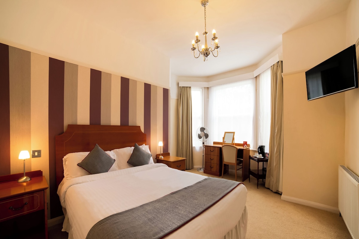 A hotel room with a large bed, a desk, a chair, a TV, and a chandelier.