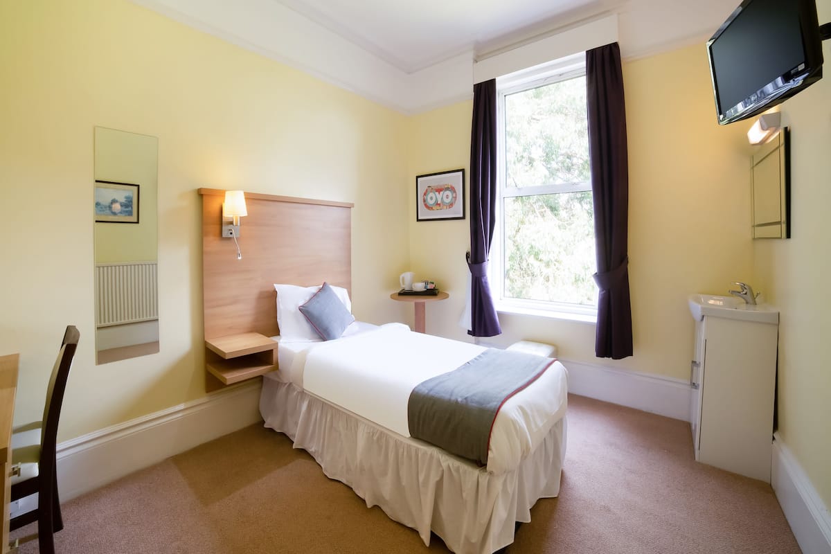 A hotel room with a bed, bedside table, lamp, window with curtains, TV mounted on the wall, and a sink.