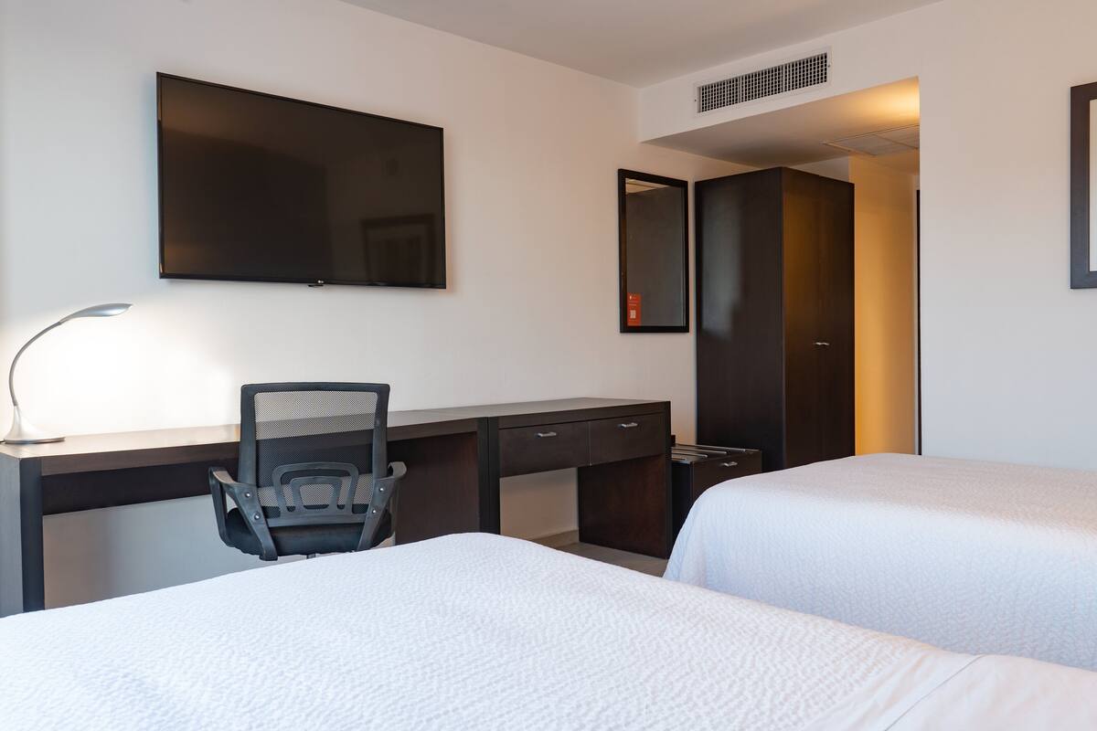 Una habitación de hotel con escritorio, dos camas, una silla, televisor y espejo.