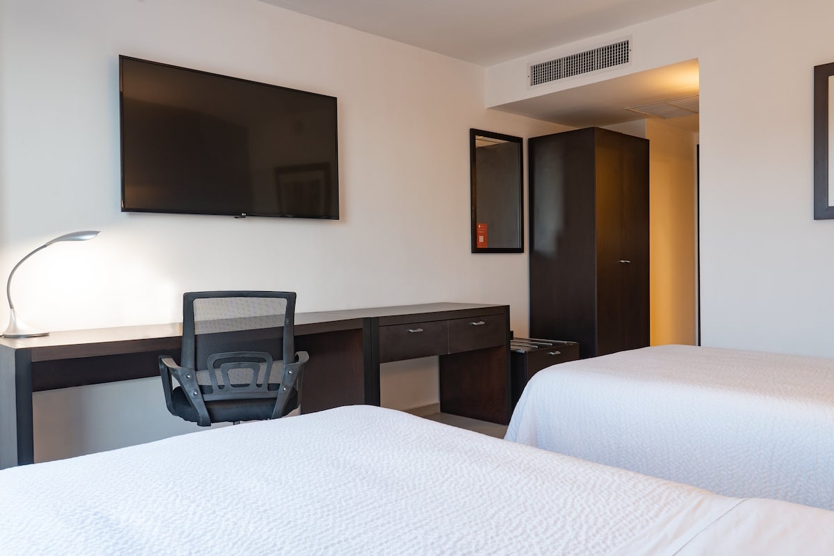 Una habitación de hotel con escritorio, dos camas, una silla, televisor y espejo.