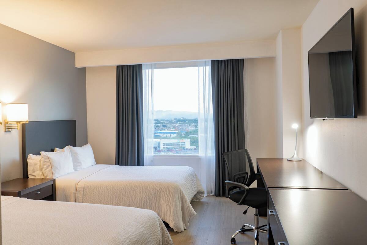 Habitación de hotel con dos camas, un escritorio, una silla, un televisor y una ventana con vistas a la ciudad.