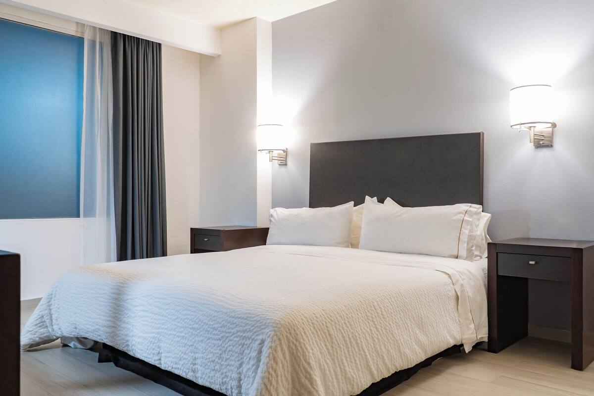 Una habitación de hotel con una cama grande, dos mesitas de noche y una ventana con cortinas.