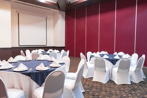 Un salón de conferencias con mesas dispuestas para una reunión, sillas blancas, una pantalla de proyector y una pared de acento roja.
