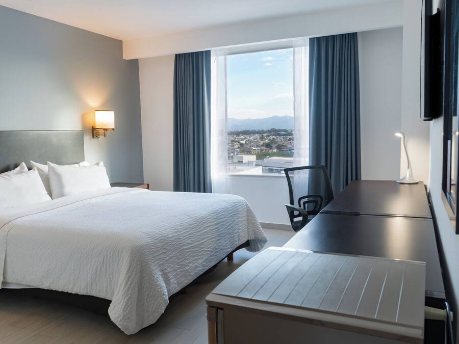 Habitación de hotel con cama, escritorio, silla, televisor y ventana con vista a la ciudad.