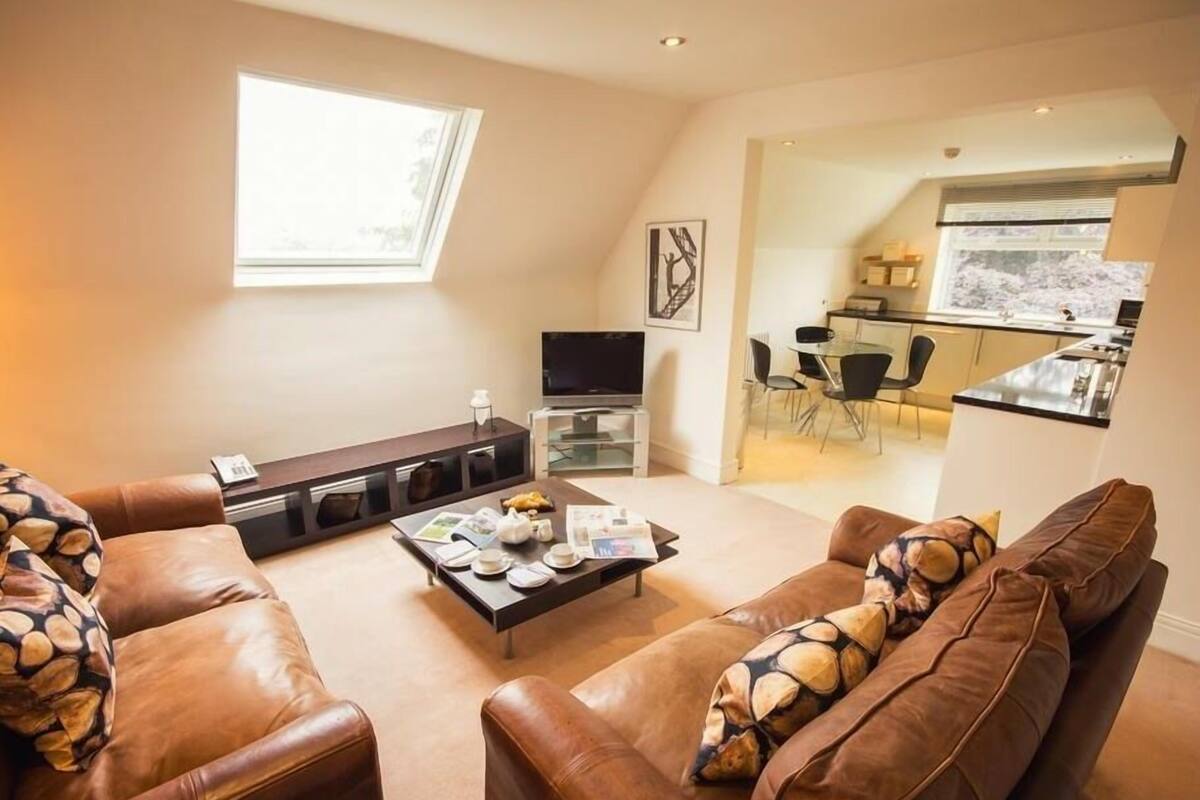 A modern living room with leather sofas, a coffee table with magazines, and a TV. A dining area with a glass table and chairs is visible in the background.