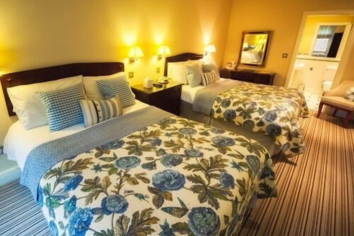 A hotel room with two beds, each with floral bedding and a headboard. There are bedside lamps, a nightstand, and a chair.