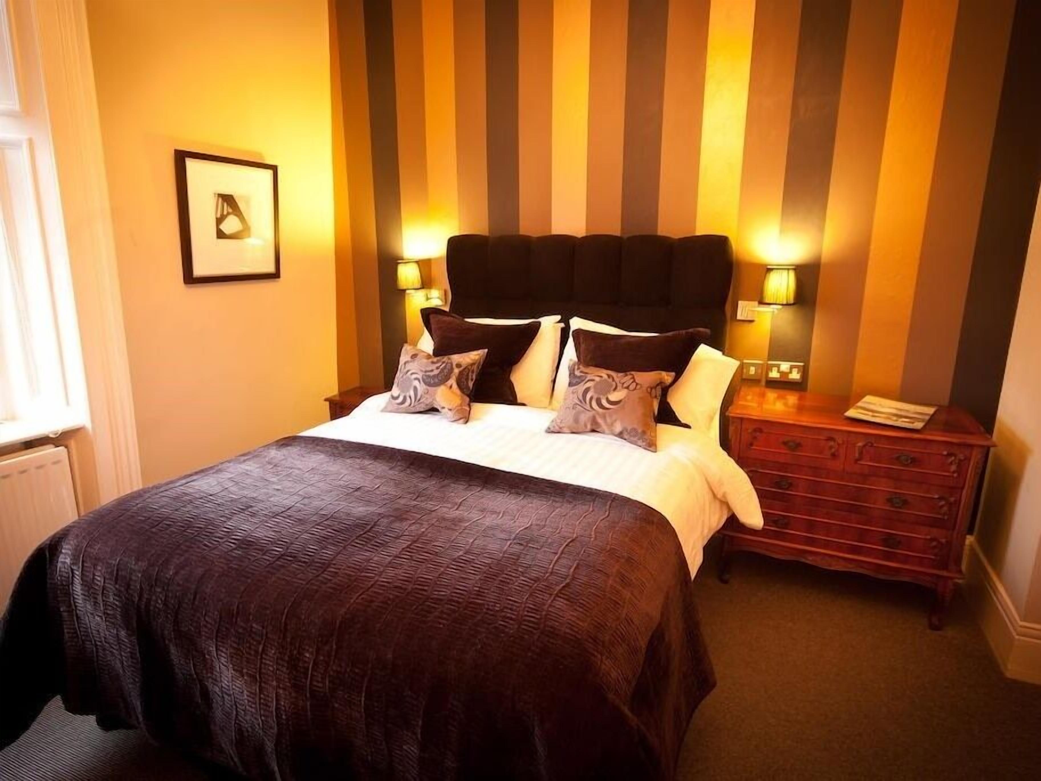 A neatly made bed with a dark brown bedspread and multiple pillows, a wooden nightstand with a lamp, and a striped wall behind the bed.