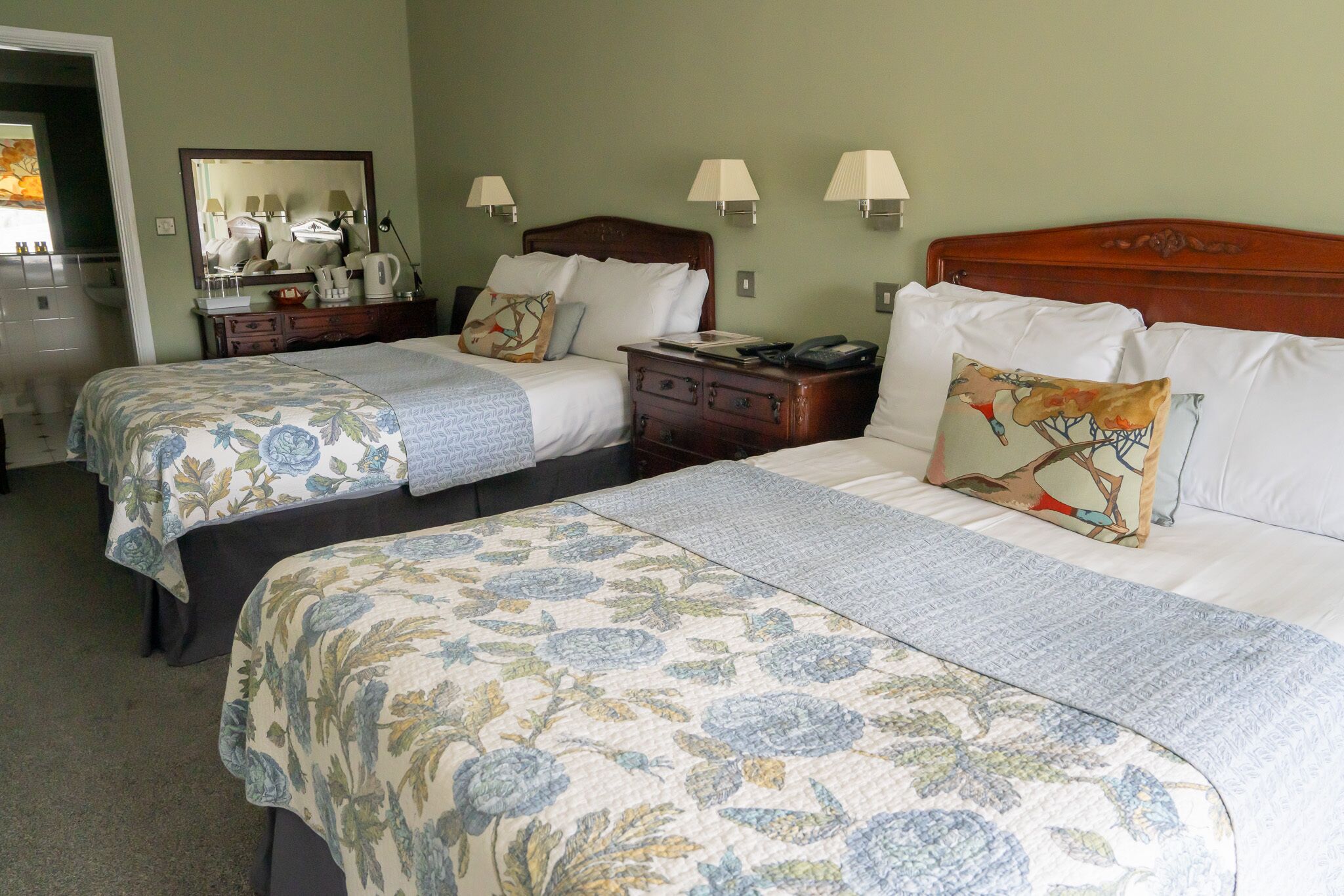 A hotel room with two beds, each with floral-patterned bedding, bedside tables with lamps, and a mirror on the wall.