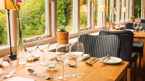 Uma sala de jantar com mesa de madeira posta para dois, taças de vidro transparentes e vista para o verde através de grandes janelas.