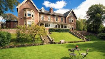 Stately mansion with a central bay window and manicured lawn, set amidst mature trees and a neatly trimmed hedge.