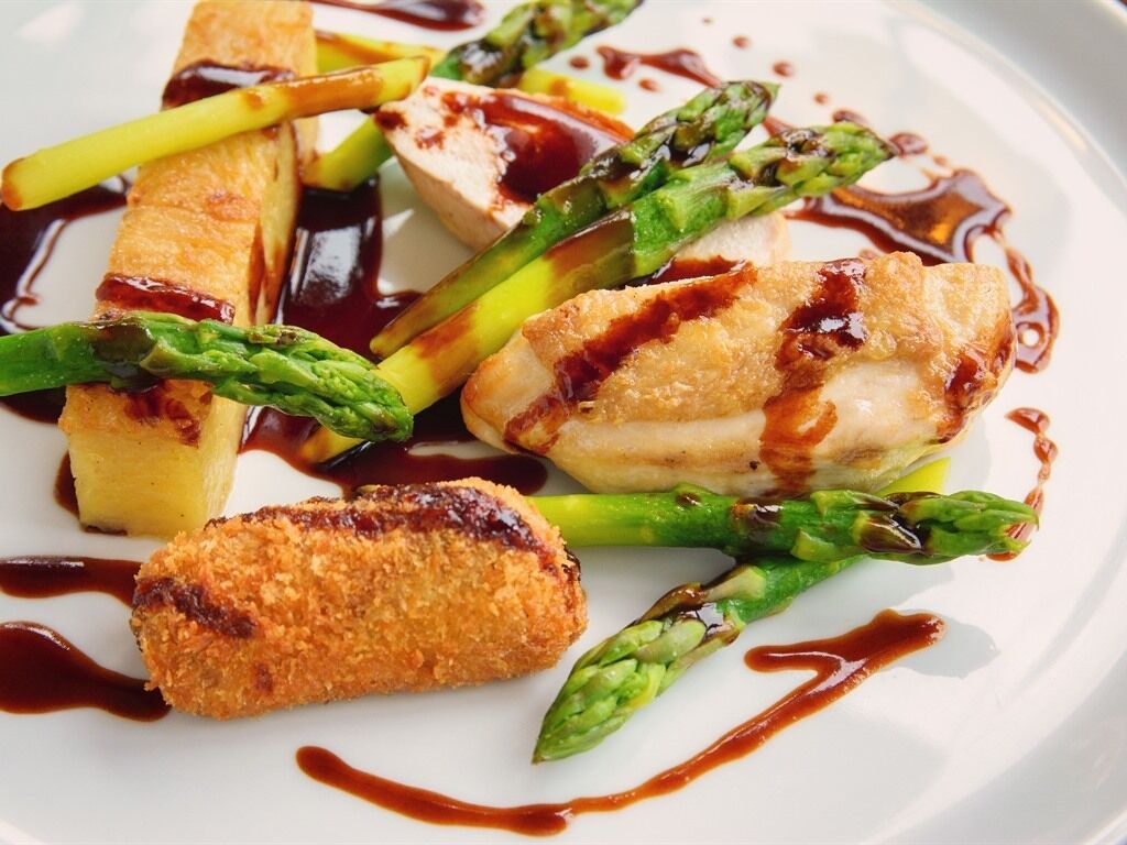 Une assiette de nourriture comprenant des asperges, un plat pané et un morceau de viande, le tout nappé de sauce.
