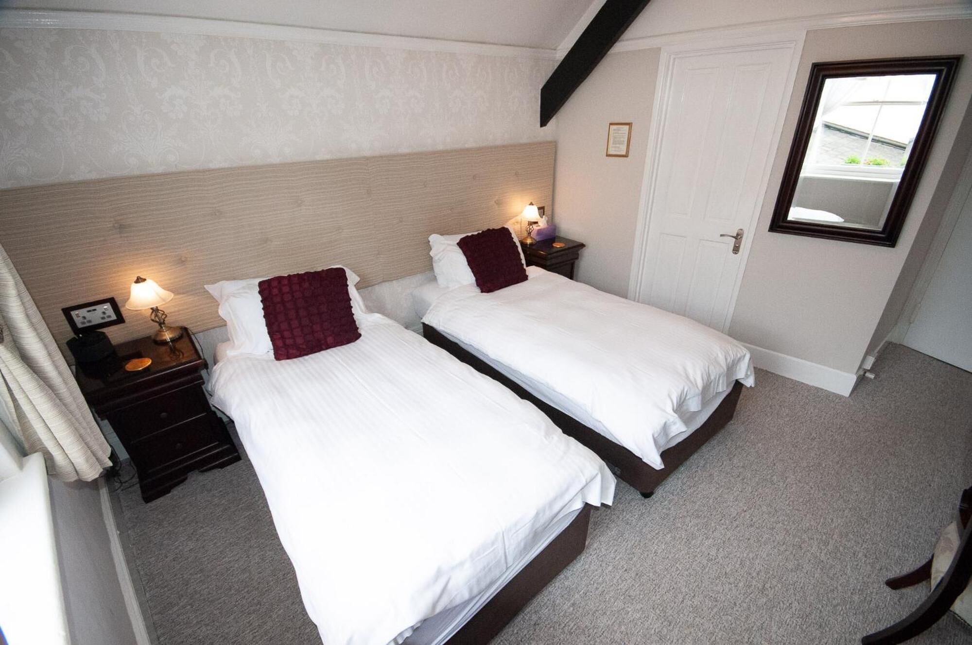 A hotel room with two single beds, a wooden headboard, bedside lamps, a small bedside table, and a window with a view.