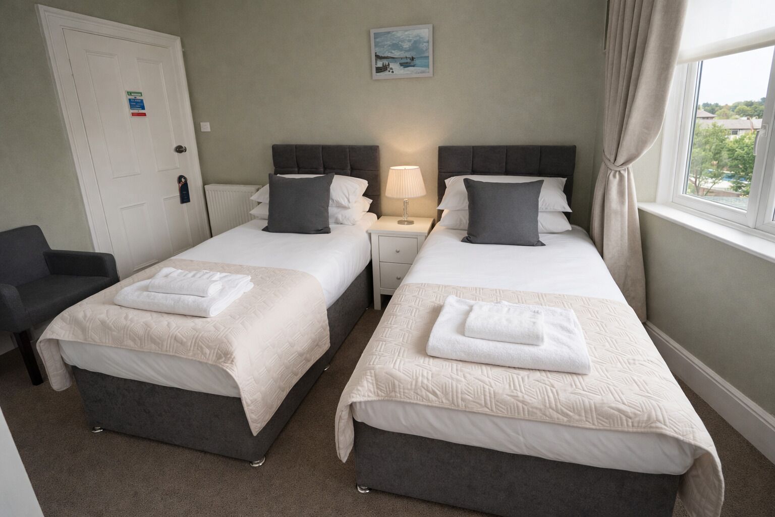A hotel room with two beds, a desk, a chair, a window with curtains, and a painting on the wall.