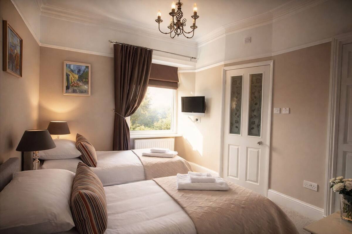A hotel room with two beds, a chandelier, a painting, a window with curtains, and a TV on the wall.