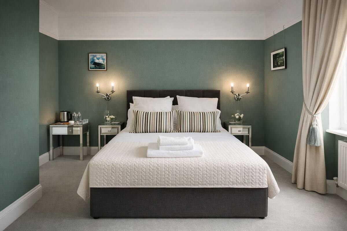 A neatly made bed with white linens and striped pillows, two bedside tables with lamps, a framed picture on the wall, and a window with curtains.