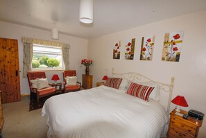 A bedroom with a large bed, two chairs, a wooden wardrobe, a window with curtains, and floral wall art. - Greyfield Farm Cottages (Bristol)