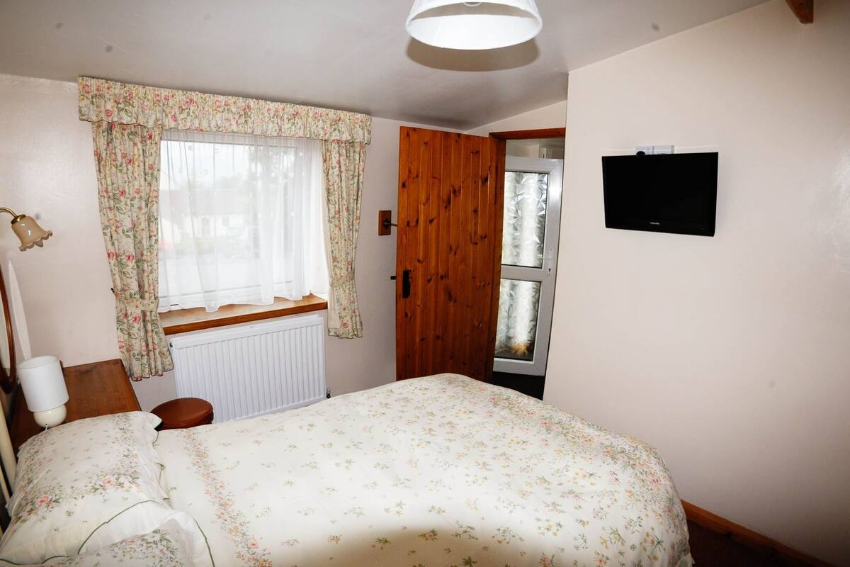 A bedroom with a bed, a window with curtains, a radiator, a wooden door, and a wall-mounted TV.
