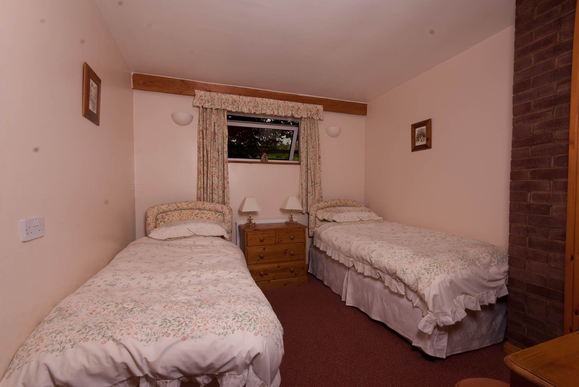 A bedroom with two single beds, a dresser, a window with curtains, and a brick wall.