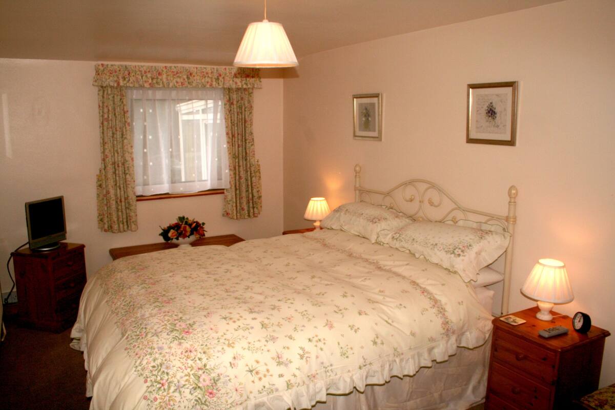 A bedroom with a bed, two bedside tables with lamps, a dresser, a television, and a window with curtains.