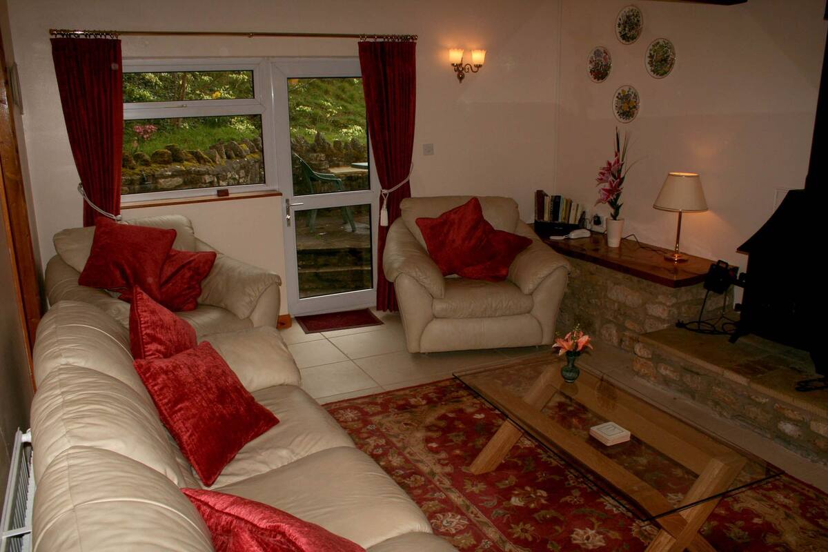 A living room with a sofa, armchair, and a coffee table. There is a window with curtains, a lamp, and a small table with a plant.