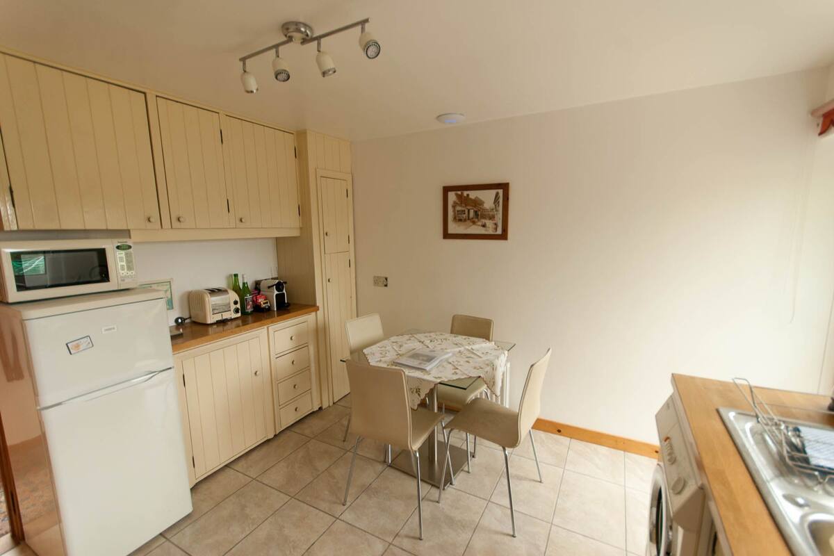 A compact kitchen with a microwave, toaster, and refrigerator. There is a dining table with chairs and a small painting on the wall.