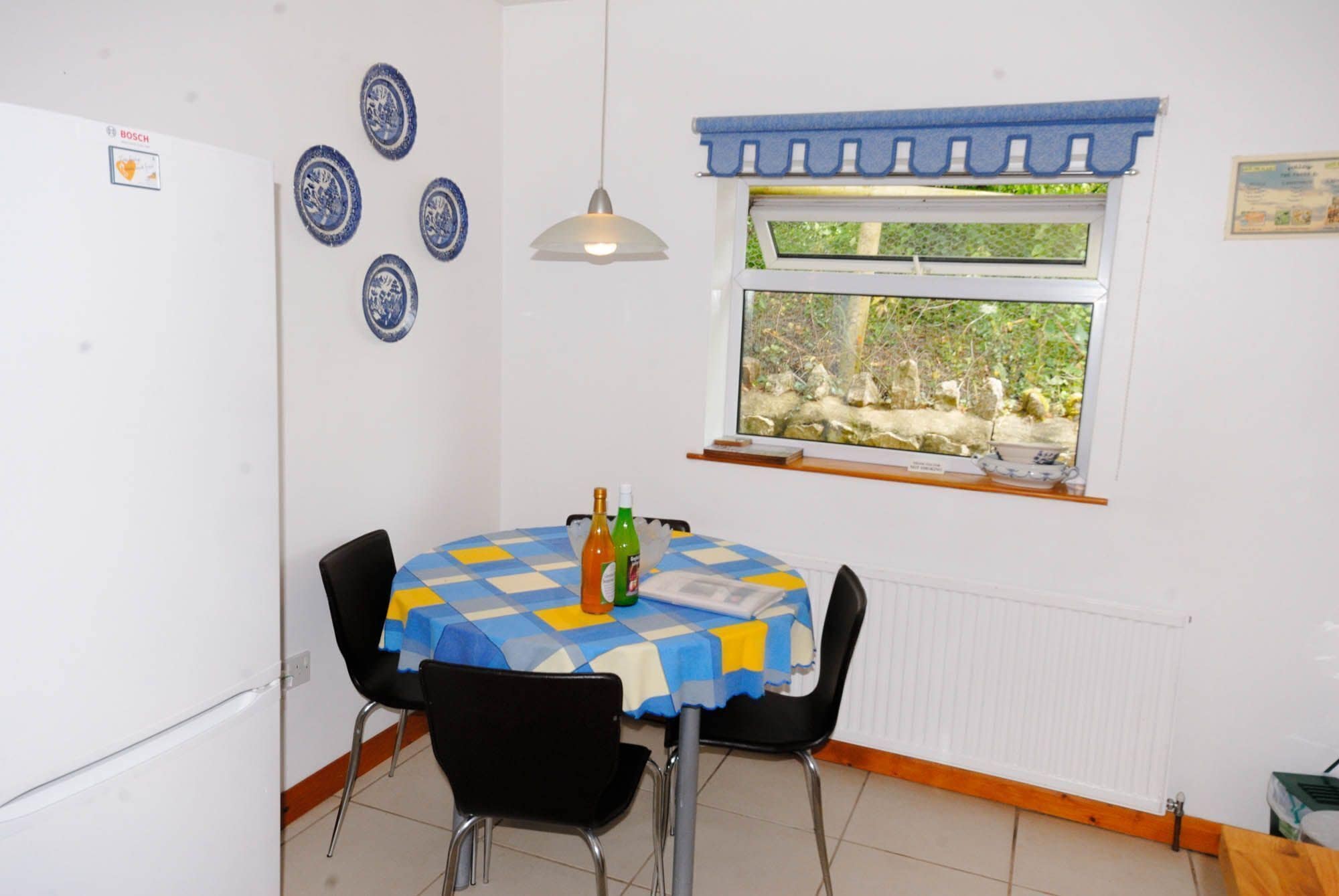 A small dining area with a round table covered by a blue and yellow checkered tablecloth, black chairs, a window with a view of greenery, and a wall-mounted Bosch refrigerator.