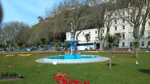 A park with a fountain, red and blue flowers, and a building in the background. - Haven House (Torquay)