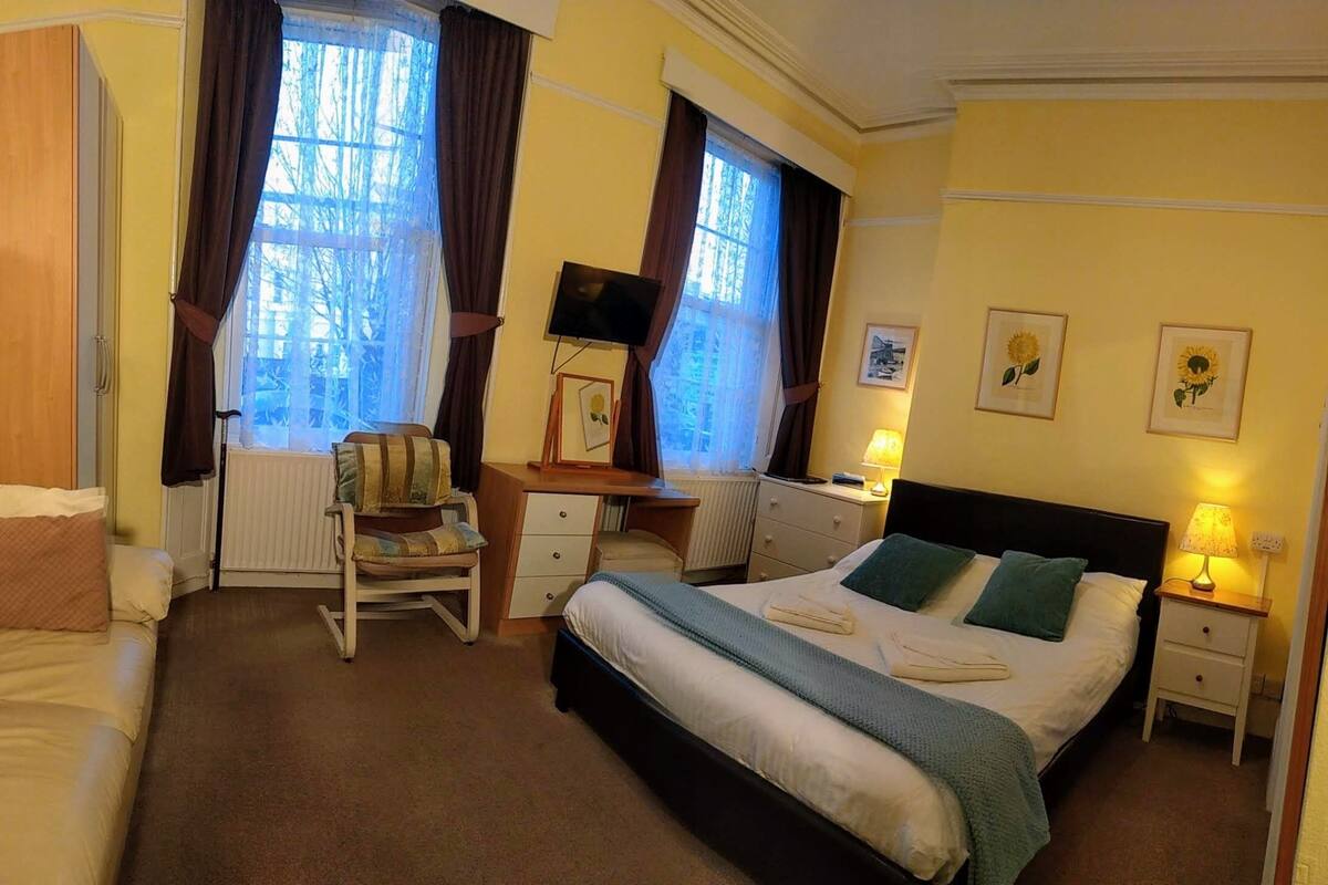 A hotel room with a bed, a desk, a chair, a radiator, and a window with curtains.