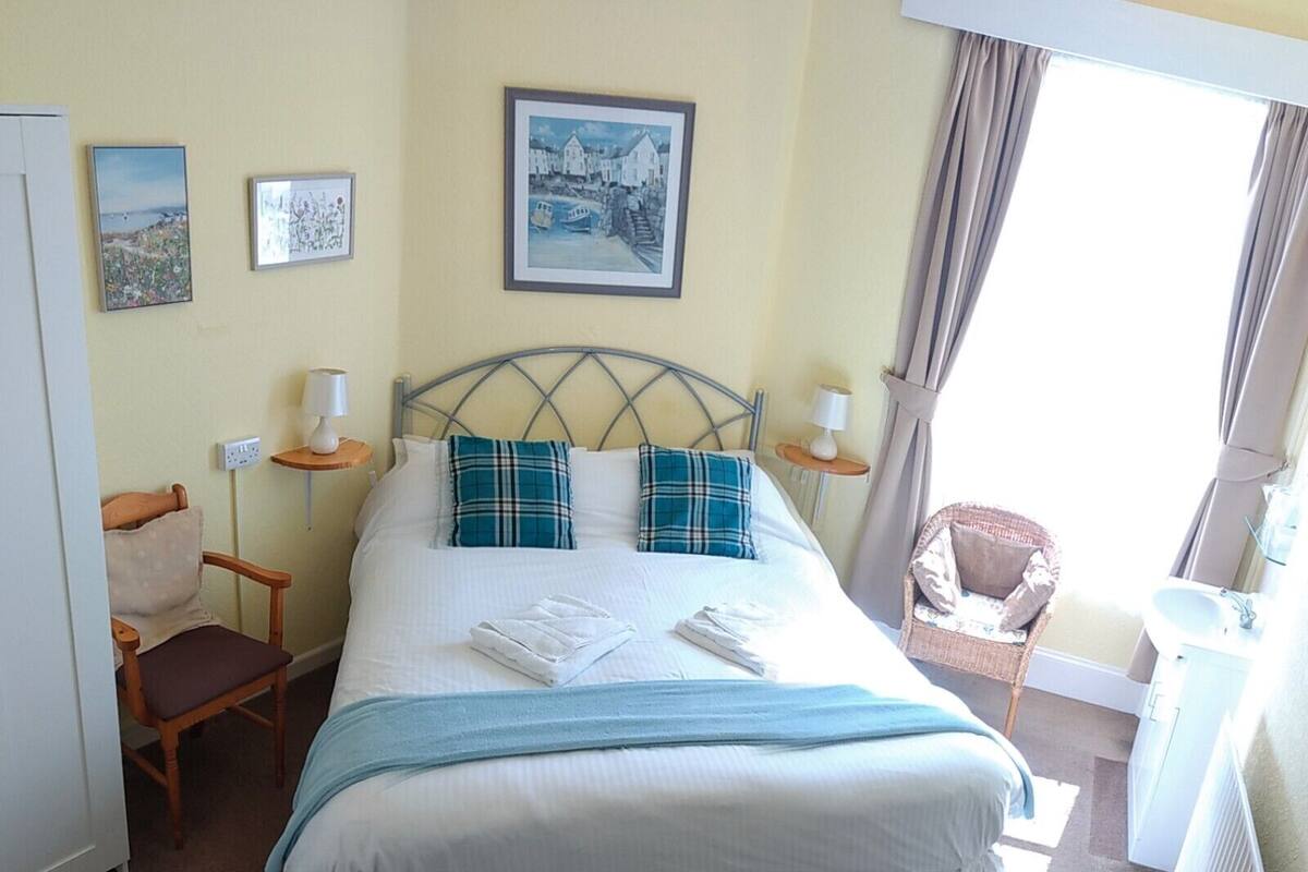 A neatly made bed with a headboard, two bedside tables with lamps, a chair, a small table, and a radiator.