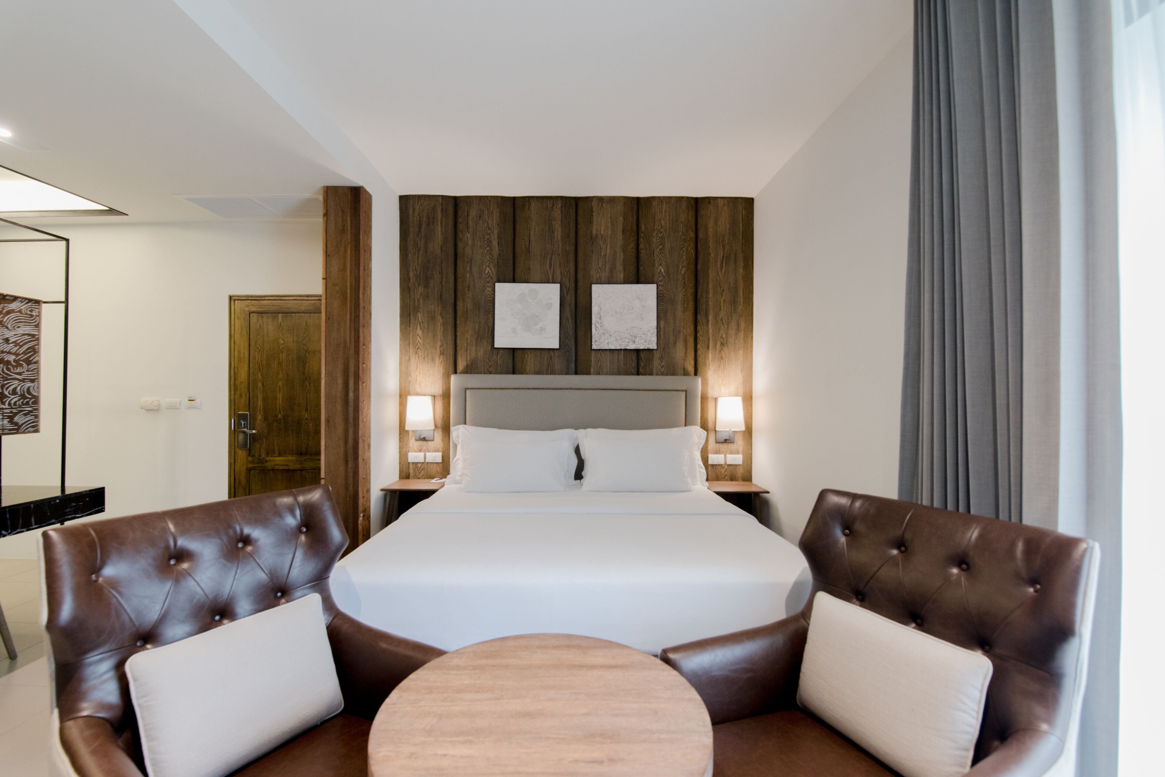 Habitación de hotel con una cama grande, dos mesitas de noche con lámparas, un cabecero de madera y dos sillones de cuero con una mesa redonda.