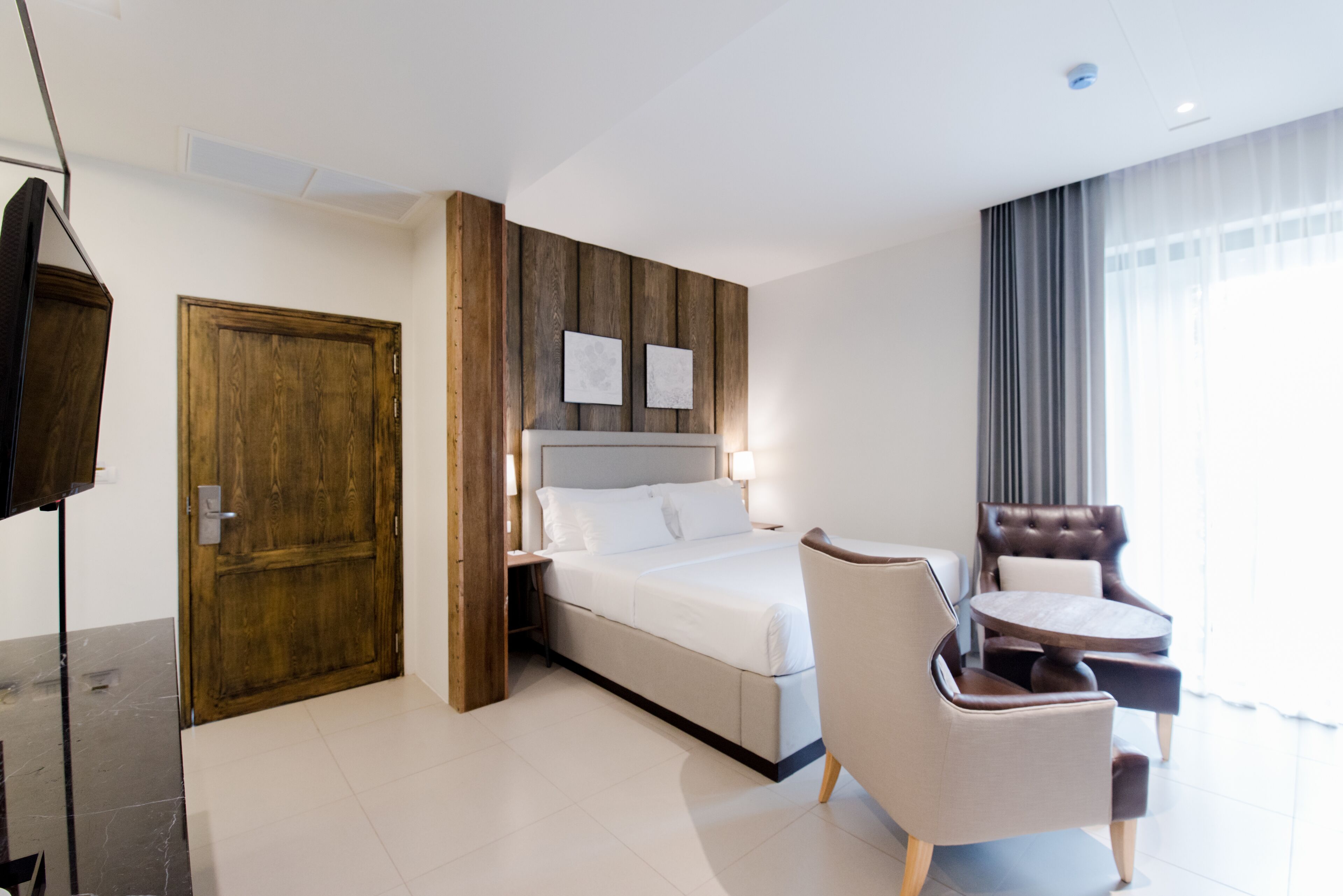A modern hotel room with a large bed, a wooden accent wall, a seating area with a round table, and a television.