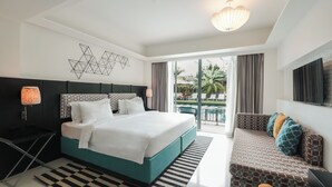 A modern hotel room with a large bed, a sofa, a TV, and a view of the outdoor pool area.