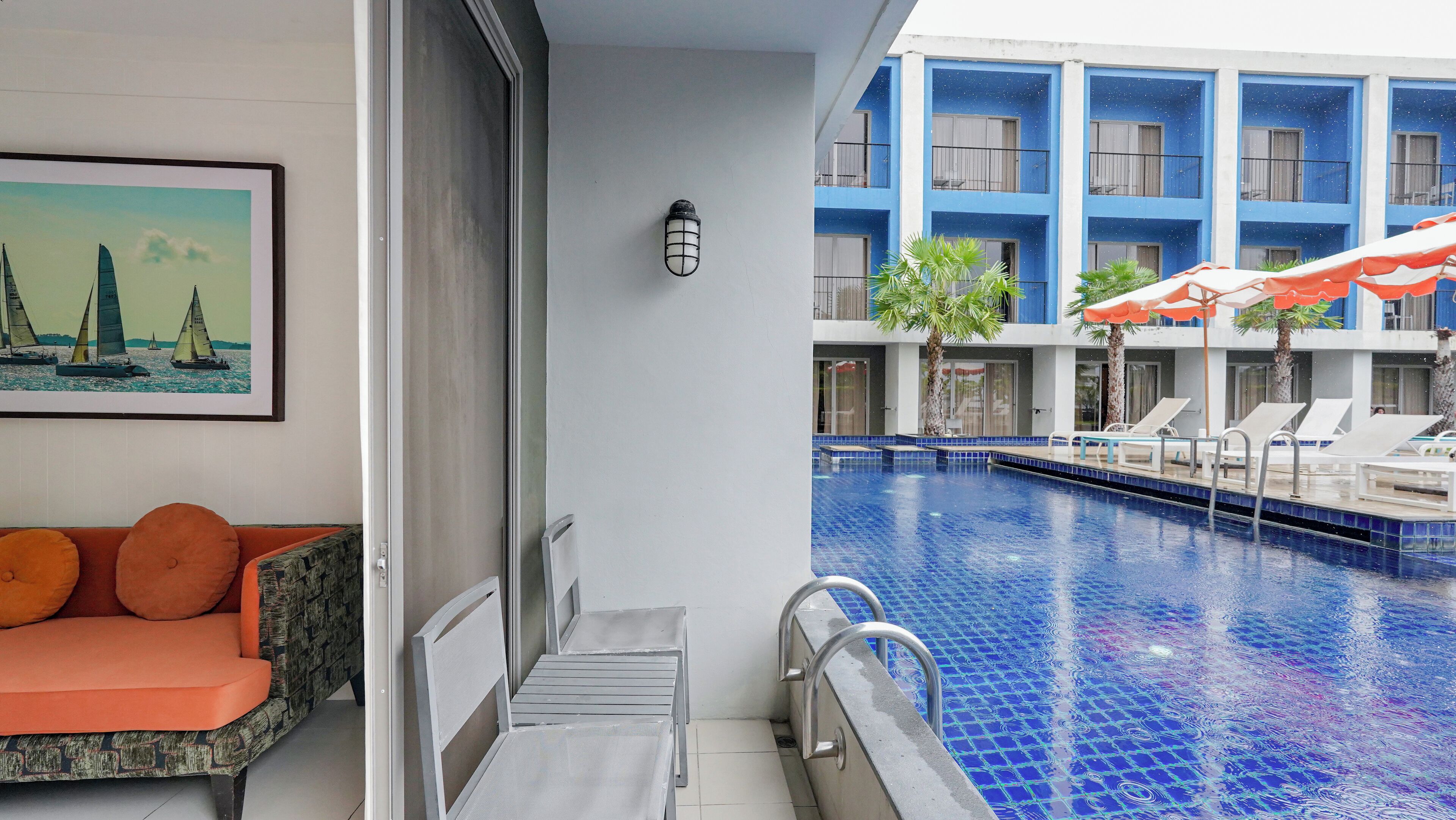 Habitación de hotel con vista a una piscina, un sofá con cojines naranja y un cuadro en la pared.