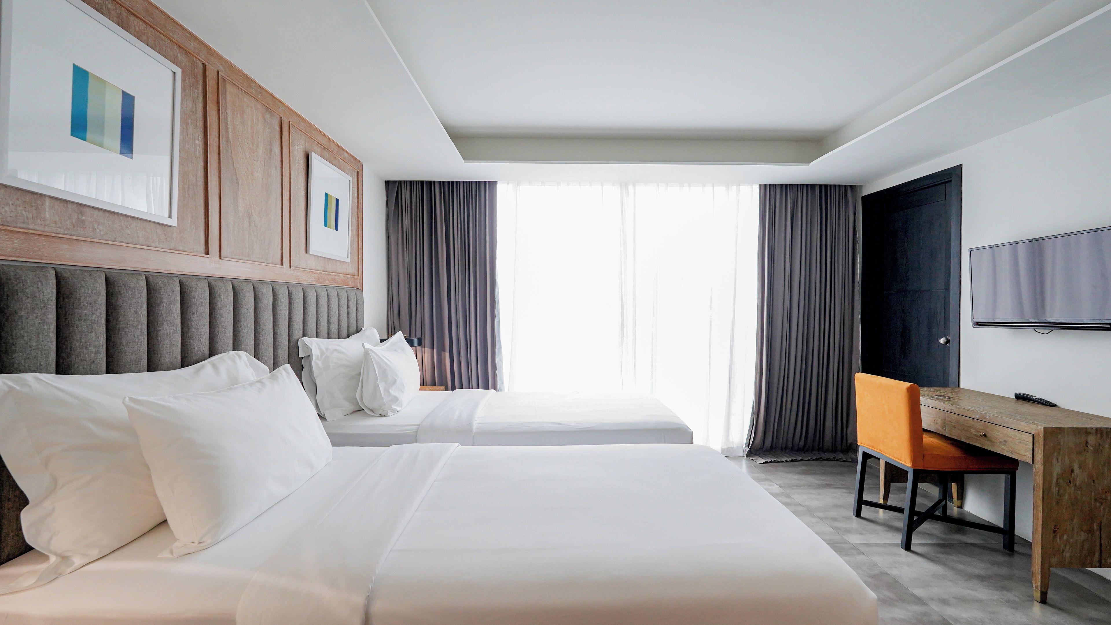 A hotel room with two beds, a desk, a chair, a TV, and a large window with curtains.