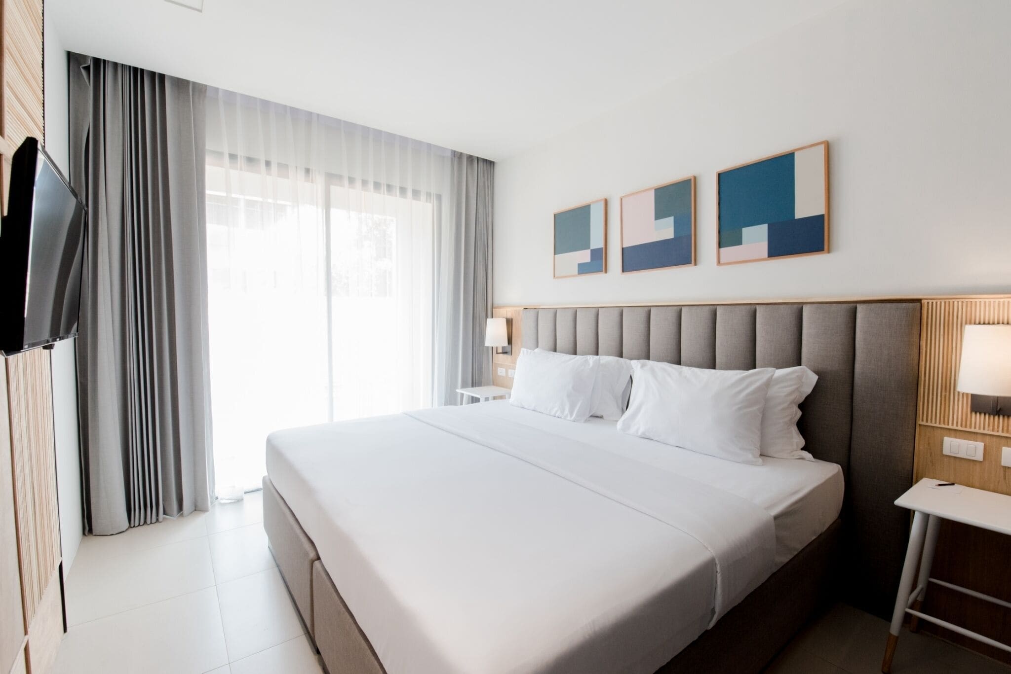 A modern hotel room with a large bed, a flat-screen TV, a bedside table, and a window with curtains.