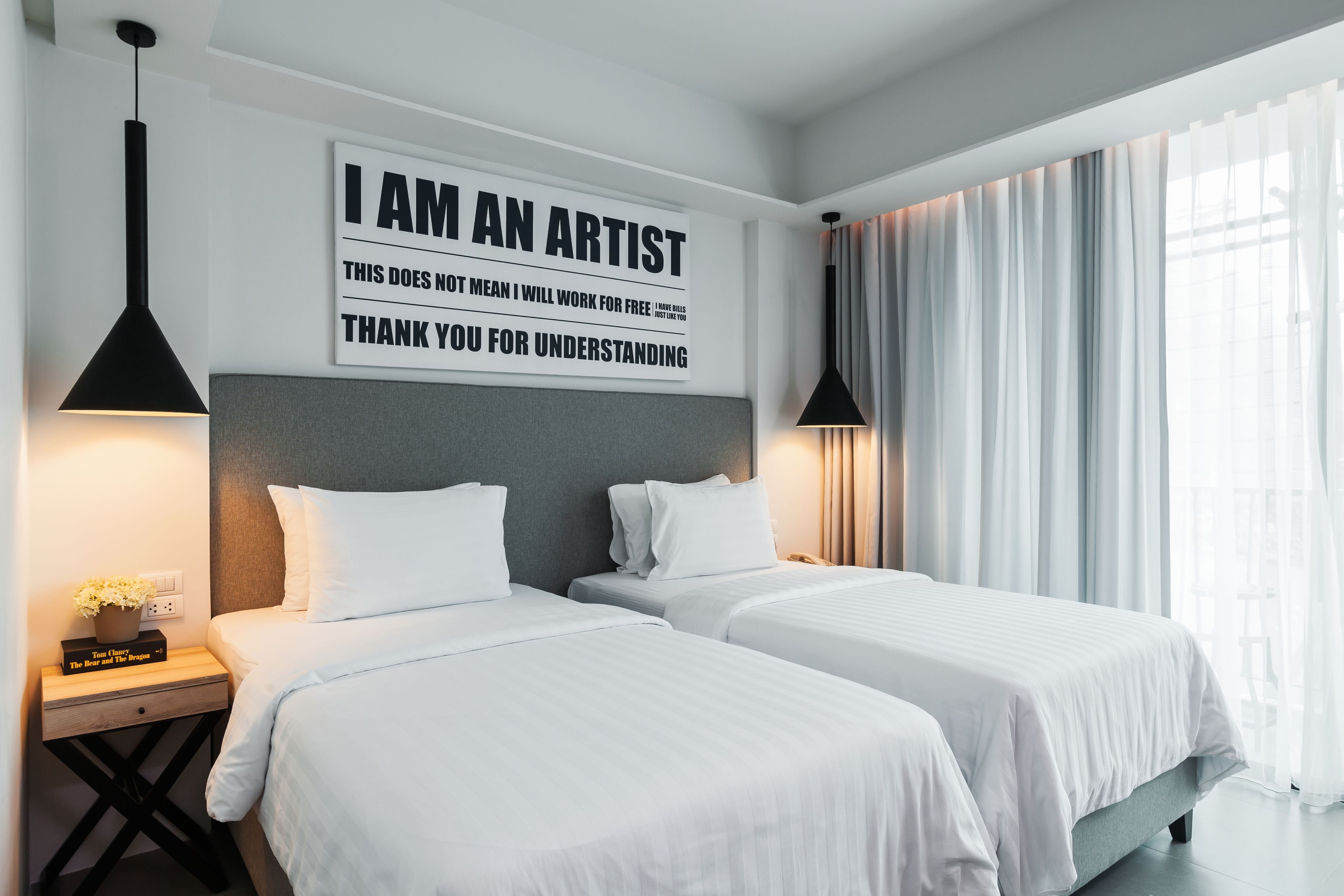 A hotel room with two beds, a bedside table with a book, a wall-mounted sign, and a window with curtains.
