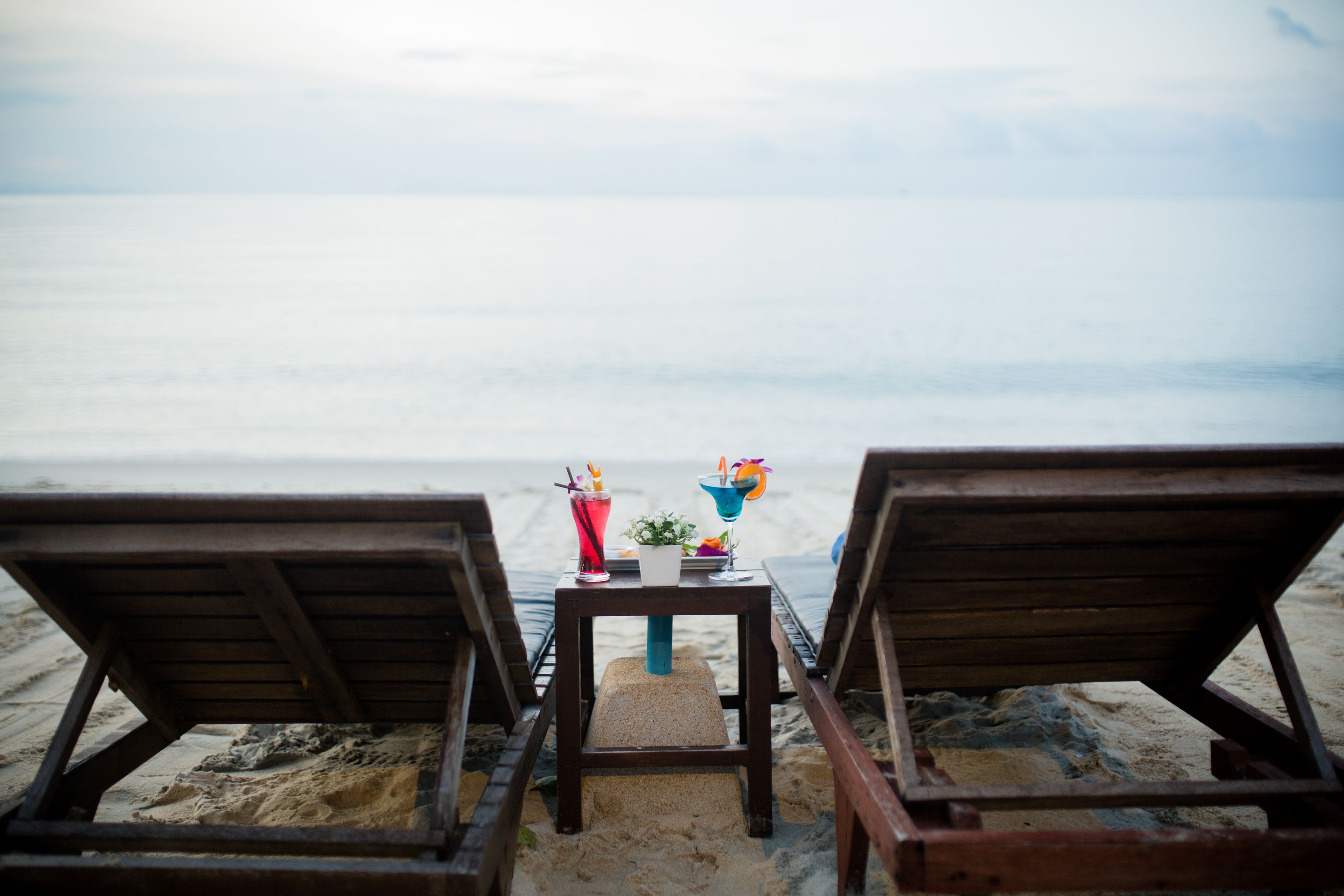 Dos sillones de madera mirando hacia una playa arenosa, con una mesa en el medio que sostiene bebidas y una pequeña planta.