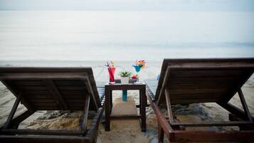 Dos sillones de madera mirando hacia una playa arenosa, con una mesa en el medio que sostiene bebidas y una pequeña planta.