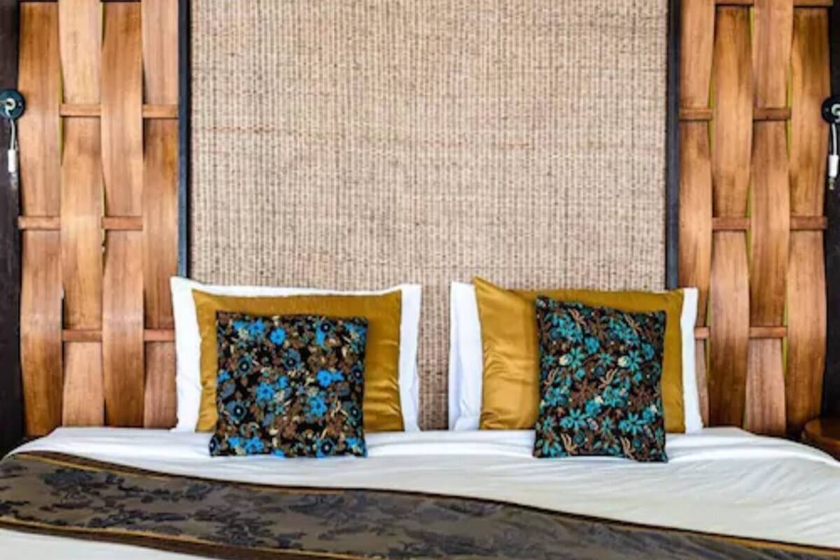 A hotel room with a woven headboard, a bed with white linens, and two decorative pillows with floral patterns.
