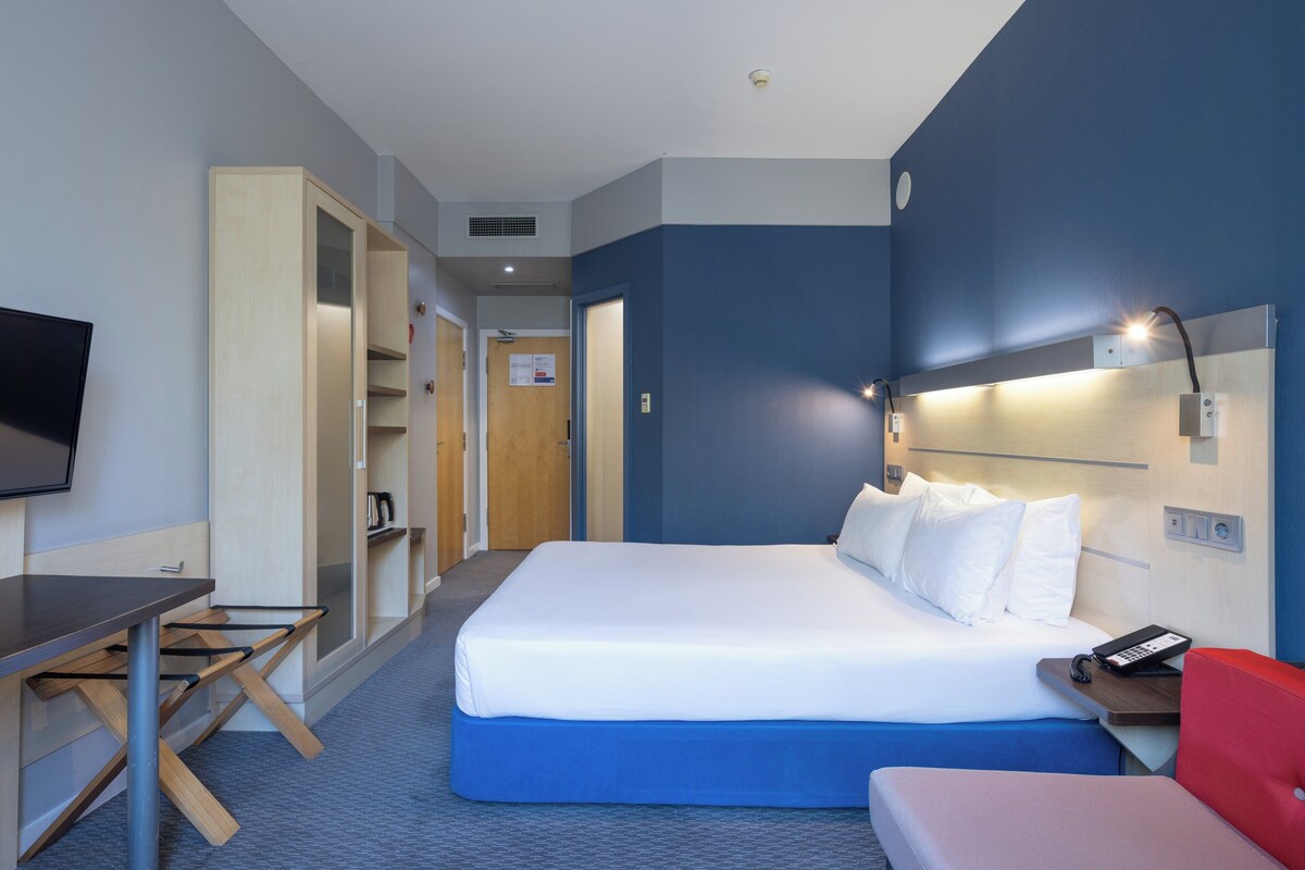 Ein Hotelzimmer mit Bett, Schreibtisch, Sessel, Fernseher und Nachttisch mit Lampe.