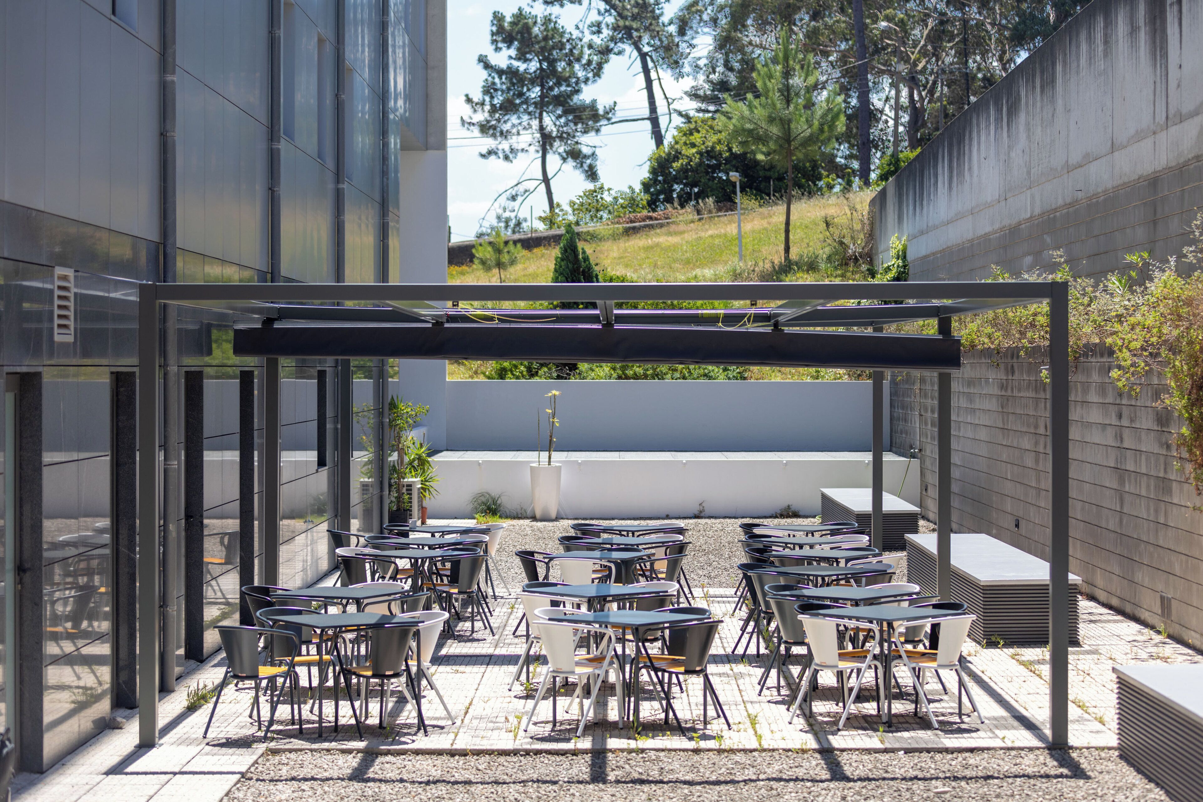 Un espace extérieur aménagé avec des tables et des chaises, une pergola et une vue sur un bâtiment et des arbres en arrière-plan.