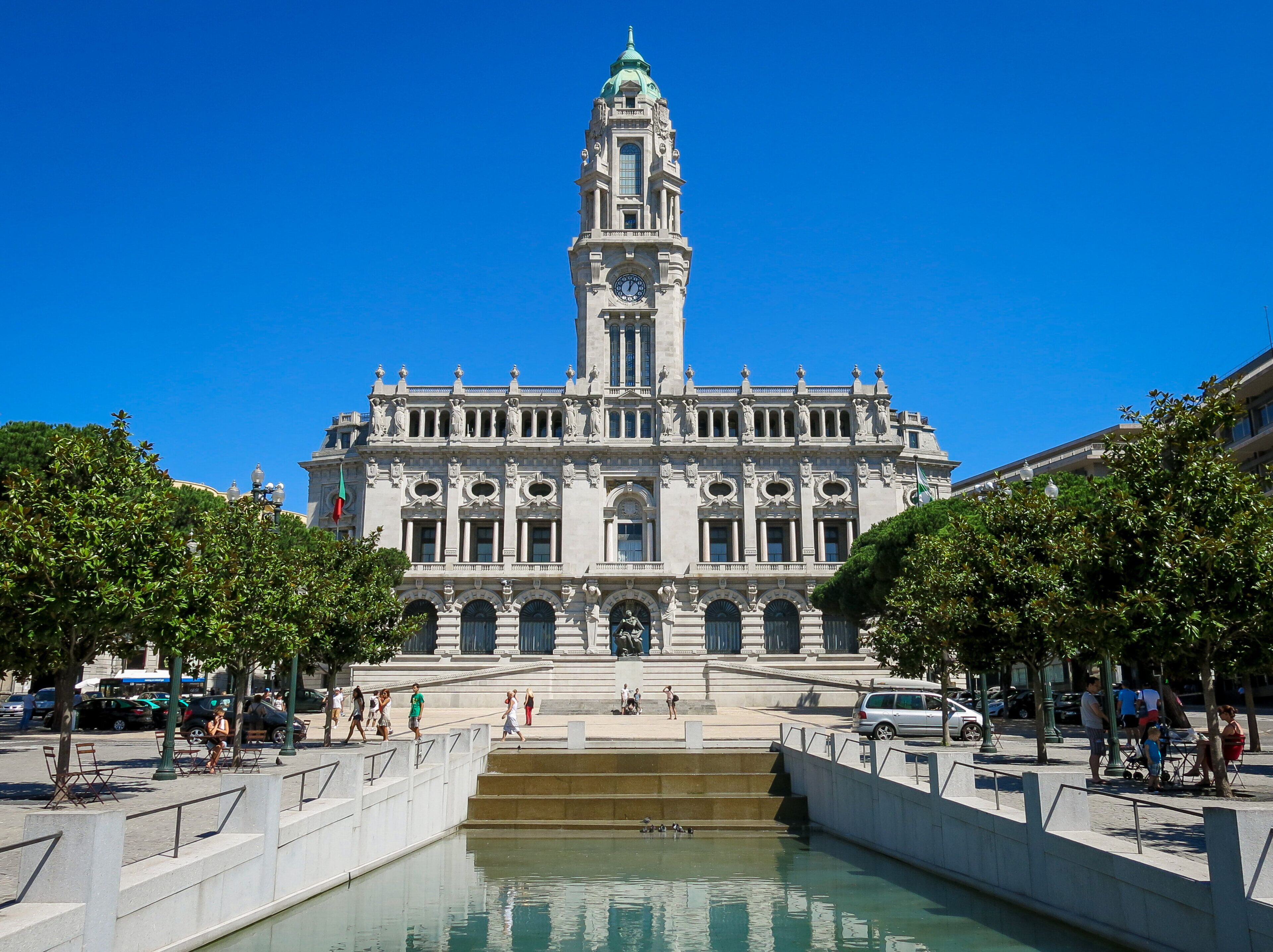 A historic building with a prominent clock tower, a central courtyard with steps leading to a fountain, and a surrounding area with trees and benches.