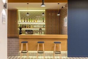 Eine Bar mit einer Holztheke, drei Barhocker und Regalen voller Flaschen und Gläsern.
