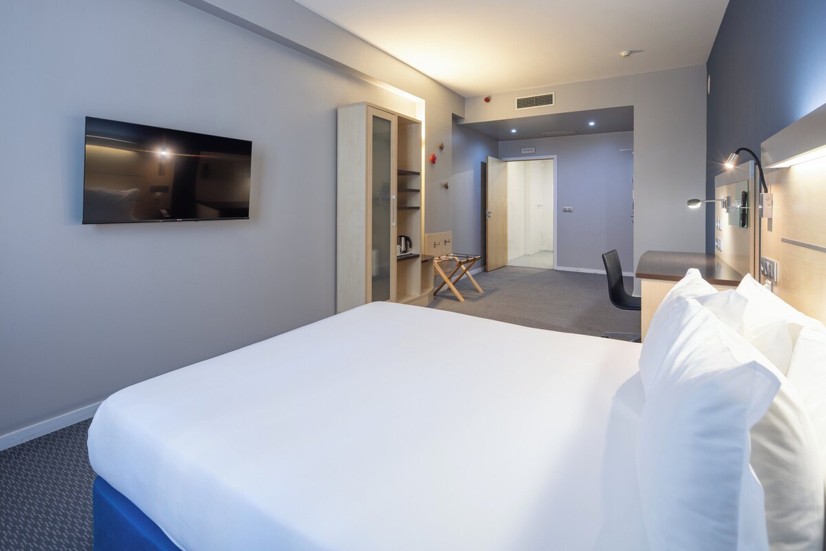 Ein modernes Hotelzimmer mit einem großen Bett, einem flachbildfernseher an der Wand, einem Schreibtisch mit Stuhl und einer kleinen Sitzecke mit Tisch und Sesseln.
