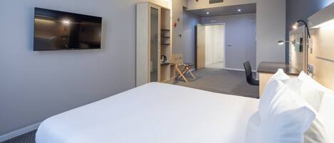 A modern hotel room with a large bed, a flat-screen TV mounted on the wall, a desk with a chair, and a small seating area with a table and chairs.