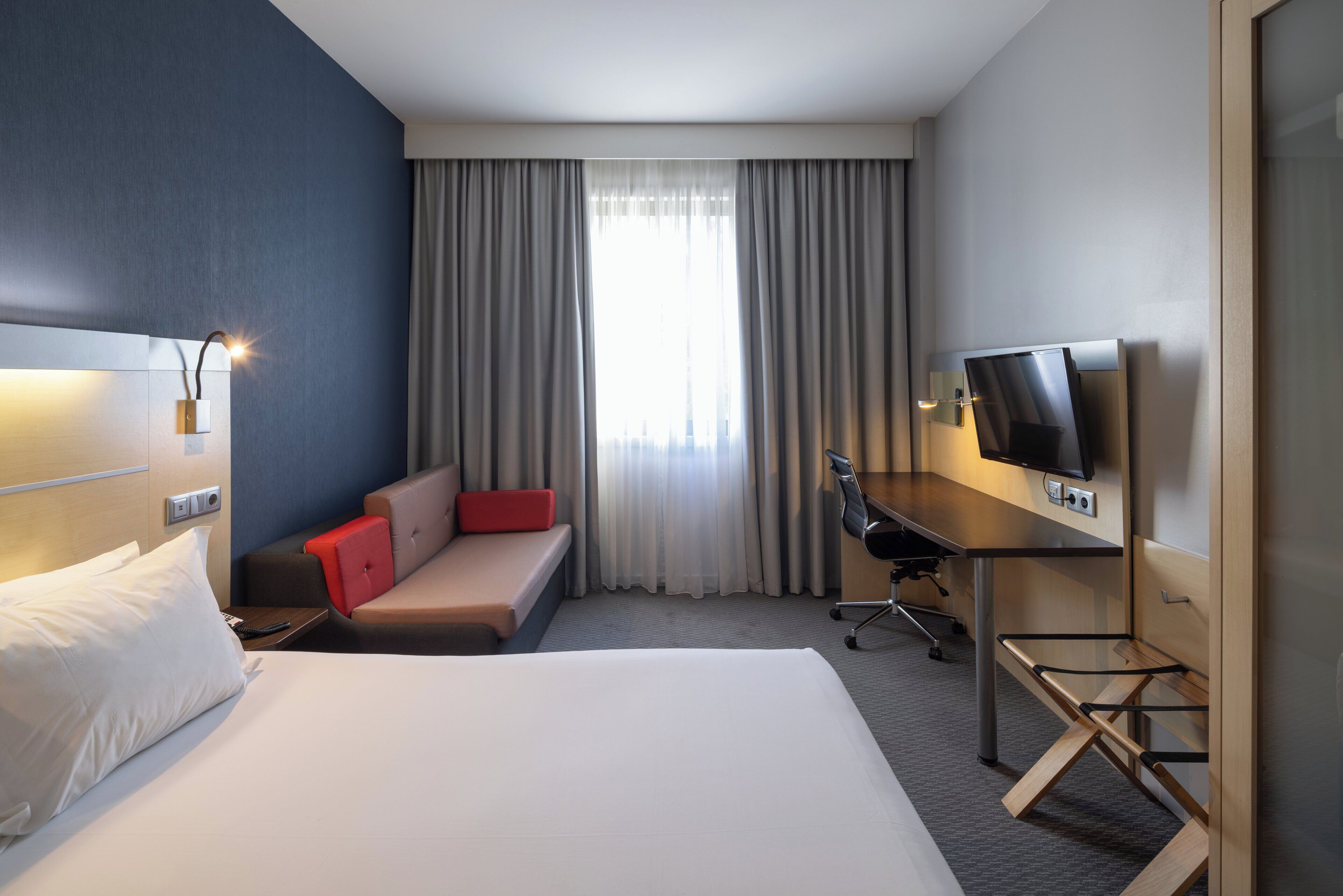 Habitación de hotel con cama, escritorio, silla, televisor y ventana con cortinas.