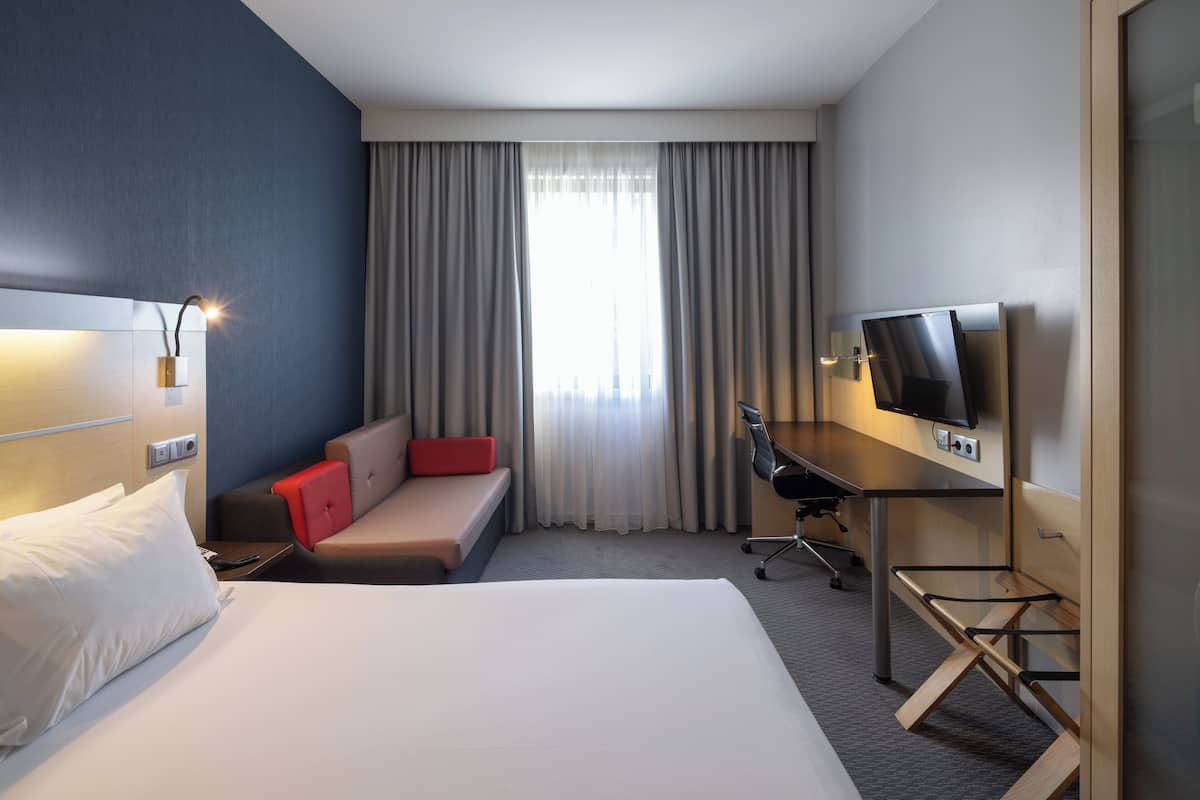 A hotel room with a bed, a desk, a chair, a TV, and a window with curtains.