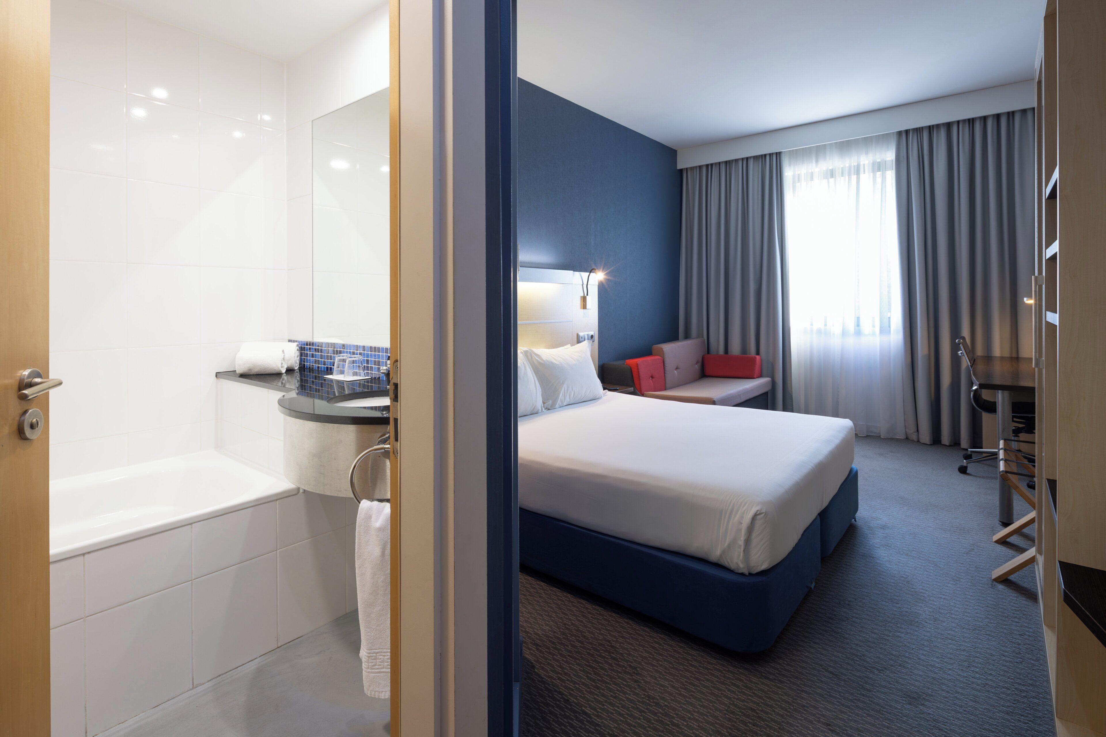 A modern hotel room with a large bed, a desk, a chair, a small table, a lamp, a window with curtains, and a bathroom with a bathtub and a towel rack.