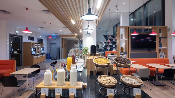 A modern breakfast area with a variety of food options, seating arrangements, and a visible kitchen area in the background. - Holiday Inn Express Porto - Exponor by IHG (Matosinhos)