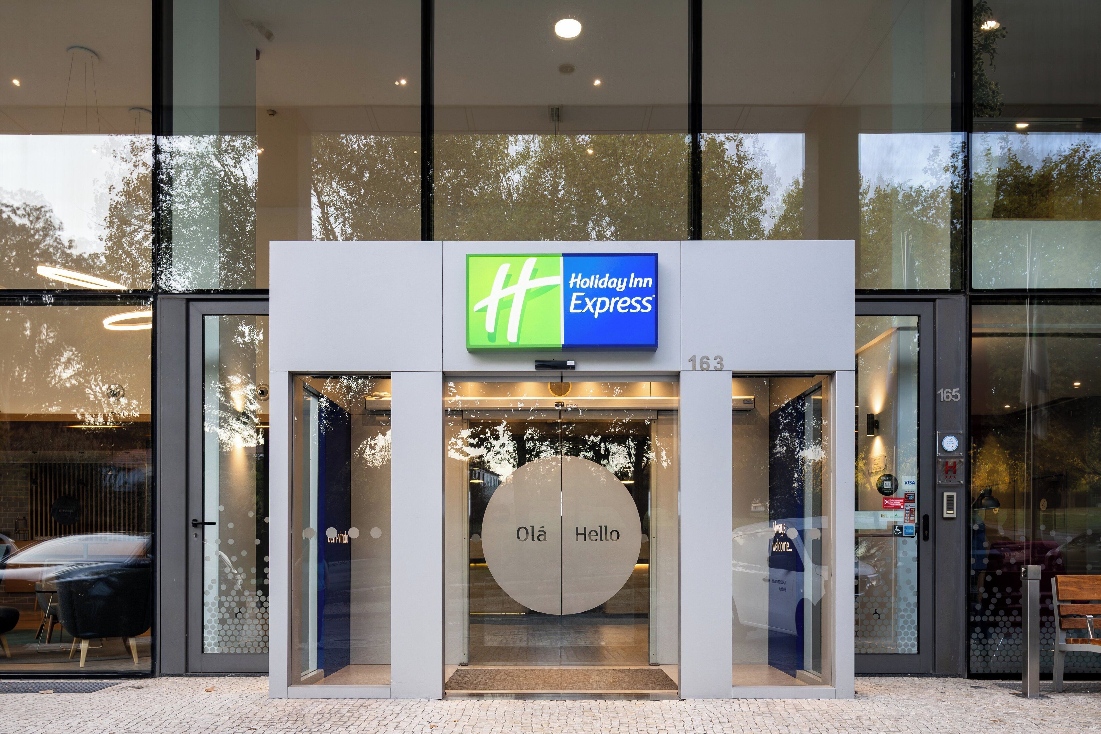 A Holiday Inn Express entrance with a glass door, a "Hello" sign, and a digital display above the door.